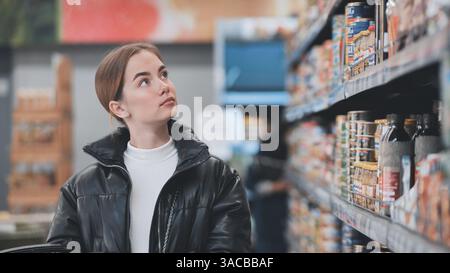 Jeune femme regardant vers le haut des produits en conserve colorés tout en faisant du shopping dans un supermarché animé, en sélectionnant soigneusement les articles pour sa liste d'épicerie Banque D'Images