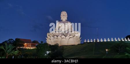 Alors que la dernière lumière du jour cède la place au crépuscule, une statue sereine et majestueuse de Bouddha blanc se dresse paisiblement au sommet d'une colline verdoyante. Banque D'Images