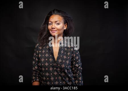 Femme souriante en kurta noir traditionnel debout sur fond noir. Jeune femme atteinte de troubles cutanés du vitiligo regardant la caméra en studio Banque D'Images