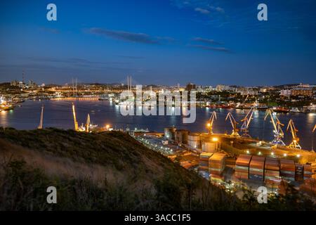 Vladivostok, Russie - 9 août 2021 : grues de chargement illuminant le port de vladivostok, russie, avec la ligne d'horizon de la ville en arrière-plan au crépuscule Banque D'Images