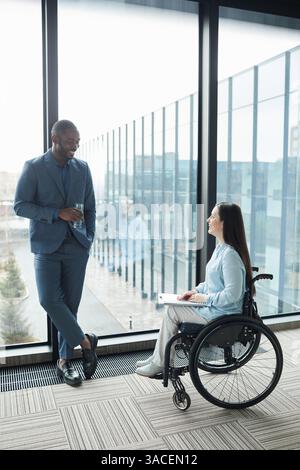 Portrait vertical pleine longueur de femme d'affaires souriante en fauteuil roulant discutant avec un collègue masculin contre la veuve dans le bureau Banque D'Images