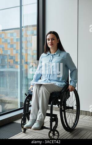 Portrait vertical pleine longueur de jeune femme d'affaires en fauteuil roulant souriant à la caméra tout en posant par fenêtre dans le bureau Banque D'Images