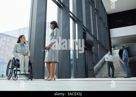 Portrait pleine longueur de jeune femme d'affaires en fauteuil roulant discutant avec une collègue féminine dans le hall du bureau, espace de copie Banque D'Images
