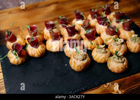 Élégants hors-d'œuvre de pâte feuilletée de la taille d'une bouchée garni de viande salée et de pâté crémeux, garnis de microgreens, disposés sur un plateau en ardoise. Banque D'Images