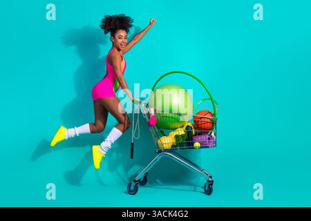 Jeune femme portant une tenue de fitness vibrante profitant d'un style de vie actif, poussant un panier rempli d'équipements sportifs Banque D'Images