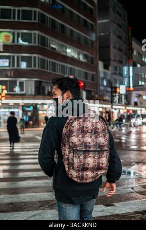 Un jeune homme portant un sweat à capuche noir et un sac à dos à motifs marche dans une rue animée de la ville la nuit. Banque D'Images