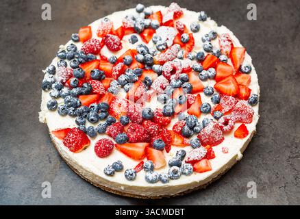 Cheesecake classique garni de fraises, framboises et bleuets, saupoudré de sucre en poudre pour un dessert festif et délicieux. Banque D'Images