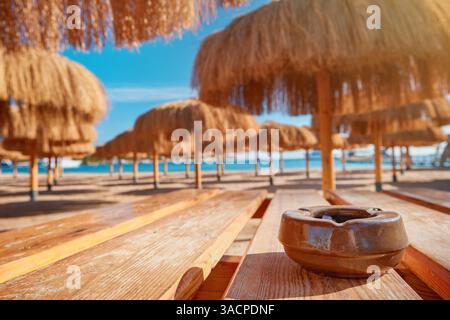 Vieux cendrier de cigarette en céramique sur fond de chaises longues, de parasols et de mer le jour ensoleillé. Concept d'habitude nocive de fumer dans les lieux ouverts. Banque D'Images