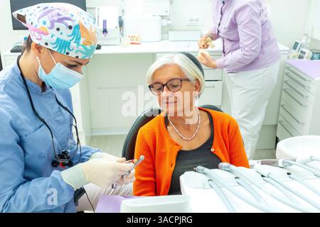 Dentiste portant un masque chirurgical et une loupe montrant une perceuse dentaire à un patient senior en clinique dentaire Banque D'Images