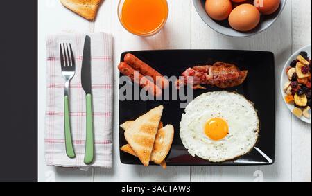 Vue de dessus d'un petit déjeuner anglais complet composé d'un œuf frit, lanières de bacon, saucisses, triangles de pain grillé, jus d'orange, oeufs et fruits secs sur une table en bois blanc, prêts à être dégustés Banque D'Images