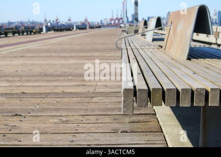 Banc de parc public avec jetée ou promenade défocalisée et grues de chargement. Passerelle en bord de mer faite de grandes planches de bois. Texture de fond de loisirs d'été. Banque D'Images