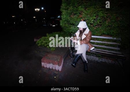 Sous la lueur des lampadaires, un individu à la mode est assis sur un banc de parc, habillé en tenue d'hiver, profitant d'un moment de calme entouré de gr luxuriants Banque D'Images