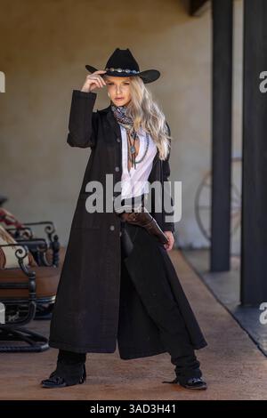 Une belle et confiante femme blonde dans une tenue de cow-boy classique se tient haute, posant avec un pistolet brillant sur sa hanche et un fusil reposant sur son shou Banque D'Images
