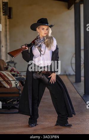 Une belle et confiante femme blonde dans une tenue de cow-boy classique se tient haute, posant avec un pistolet brillant sur sa hanche et un fusil reposant sur son shou Banque D'Images