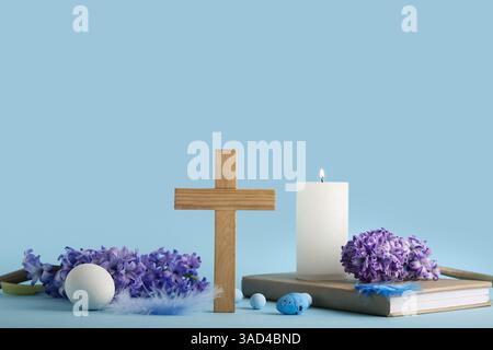 Croix en bois avec bougie allumée, livre, fleurs de lilas et œufs peints sur fond bleu. Fête de Pâques Banque D'Images