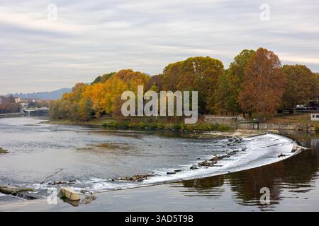 Le fleuve po à Turin, Italie, avec un petit barrage créant des cascades douces. Feuillage d'automne coloré tapisse les berges sous un ciel couvert. Banque D'Images