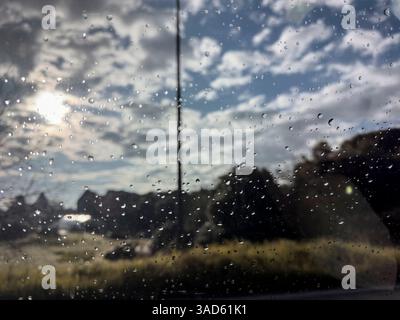 Les gouttes de pluie accrochées à une fenêtre de voiture obscurcissent la vue sur le paysage au-delà, créant une scène floue et impressionniste par temps nuageux Banque D'Images