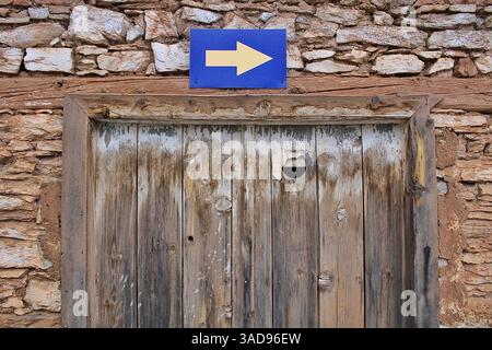 Vieille porte en bois sous un signe de flèche bleue sur un mur de pierre rustique. Parfait pour les thèmes du voyage, de la navigation ou de l'architecture traditionnelle. Banque D'Images