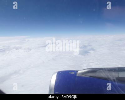 Vue aérienne depuis une fenêtre d'avion montrant des nuages blancs moelleux et un ciel bleu. L'aile de l'avion est partiellement visible au premier plan, avec du soleil Banque D'Images