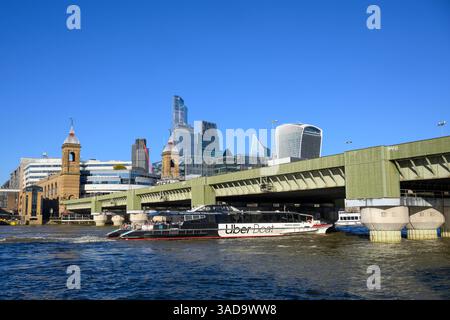 Un Uber Thames Clipper passe sous le pont ferroviaire et la gare de Cannon Street, avec les gratte-ciel du quartier financier de la City of London en t Banque D'Images