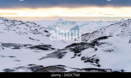 Un paysage hivernal à couper le souffle avec des rochers enneigés au premier plan, menant à une vaste étendue glacée avec d'imposants icebergs au loin. Banque D'Images