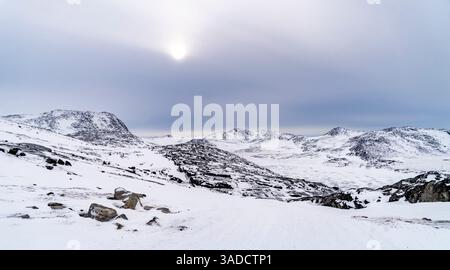 Un paysage hivernal serein avec des montagnes enneigées et un ciel nuageux. Le soleil est partiellement visible, projetant une lumière douce sur le terrain enneigé Banque D'Images