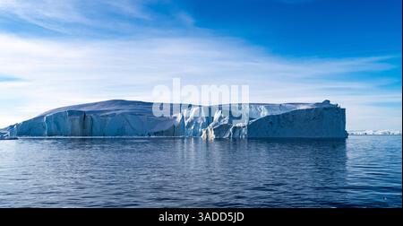 Une vue imprenable sur un grand iceberg flottant dans des eaux calmes sous un ciel bleu clair. L'iceberg présente des arêtes vives et dentelées et reflète magnifiquement i Banque D'Images