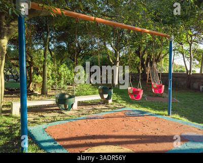 Quatre balançoires pour tout-petits dans un parc calme avec des arbres et la lumière chaude du soleil du soir, pas de personnes présentes Banque D'Images