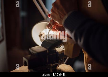 Luthier sculpte méticuleusement un morceau de bois, serré dans un étau, à l'aide d'une gouge, sous la lueur chaude d'une lampe, mettant en valeur l'artisanat impliqué dans Banque D'Images