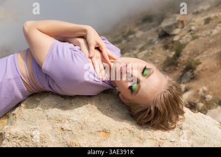 Mannequin de mode féminin avec les yeux fermés se trouve sur le dos sur le rocher dans les montagnes, place les deux paumes sur son cou et détendu à l'extérieur. Sensualité femme avec Mak Banque D'Images