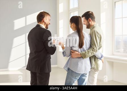 Agent immobilier souriant en costume montrant la disposition de l'appartement à un couple heureux dans une pièce vide lumineuse. Banque D'Images