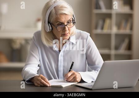 Femme d'affaires âgée dans le casque regarder webinaire sur ordinateur portable prendre des notes Banque D'Images