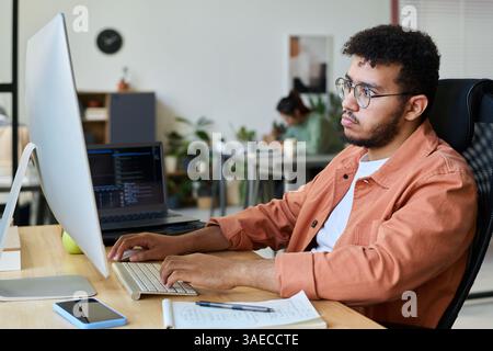 Sérieux jeune développeur ou concepteur de logiciel masculin appuyant sur les touches du clavier de l'ordinateur et regardant à travers l'information en ligne Banque D'Images