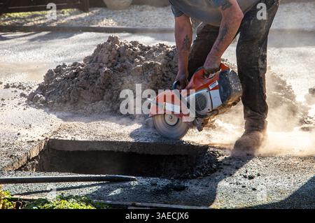 Ouvrier de la construction utilisant une scie électrique pour couper à travers une allée percée pour accéder à un tuyau de buste pour réparation par une journée ensoleillée Banque D'Images