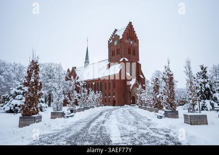Un chemin enneigé guide le chemin vers une église en briques rouges frappante, nichée parmi les arbres glacés dans un village d'hiver serein à Hassleholm, en Suède. Banque D'Images