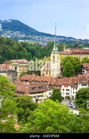 Berne, Suisse, 30 mai 2024. Cette image capture la superbe ligne d'horizon de Berne, en Suisse, avec l'historique Nydeggkirche et Berne Minster. La scène souligne le charme médiéval de la ville, avec ses toits en tuiles rouges, sa végétation luxuriante et ses tours d'église emblématiques dressées contre un ciel bleu vibrant. Banque D'Images