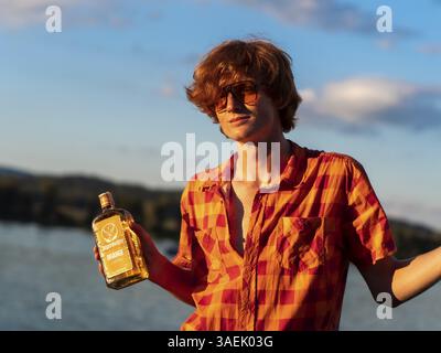 Jeune homme debout près du lac, tenant une bouteille d'alcool dans ses mains, liqueur aux herbes Jaegermeister, cheveux rouges, chemise orange, portrait Banque D'Images