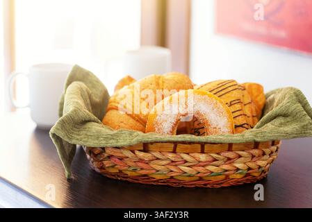 Panier tissé rempli d'un assortiment de pâtisseries fraîches, y compris des croissants, beignets et pain au chocolat, se trouve sur une table à côté d'une tasse blanche, suggérant une scène de petit déjeuner délicieux Banque D'Images