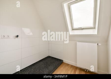 Cette image met en valeur une salle de bains contemporaine avec un plafond incliné, des carreaux blancs et un chauffage élégant. Le design minimaliste met l'accent sur la lumière et l'espace. Banque D'Images