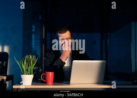 Jeune homme d'affaires travaillant tard dans la nuit dans un bureau faiblement éclairé, regardant son ordinateur portable avec une expression fatiguée. Concept de surmenage, longues heures, fatigue Banque D'Images