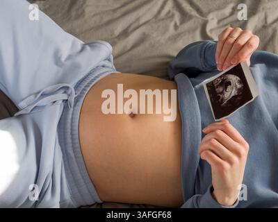 Femme enceinte reposant confortablement dans son lit, portant des vêtements de détente bleus doux, serrant tendrement l'image échographique contre le ventre de la femme enceinte, embrassant Banque D'Images