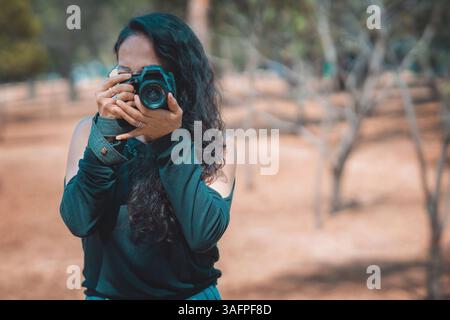 Femme photographe tenant un appareil photo professionnel, prend une photo dans le parc en se concentrant sur quelque chose, le concept de jour du photographe. Banque D'Images
