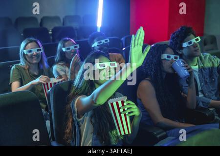 Groupe d'amis profitant d'une expérience de film 3D passionnante dans le cinéma, grignoter sur pop-corn et lever les mains excitamment dans le plaisir Banque D'Images