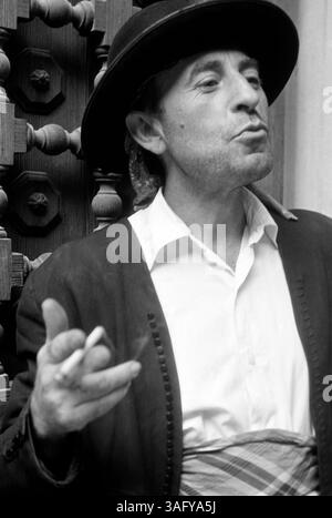 Ein mallorquinischer Mann gestikuliert mit einer Zigarette in der rechten hand beim Sprechen, er trägt eine lokale Tracht, die aus einem schwarzen Hut, einem weißen Hemd mit karierter Bauchschärpe und einer kragenlosen Jacke mit eng Rücken Knopfleiste besteht, hinter seinem bestückter ist eine ortstypische Schnitzarbeit, möglicherweise Tür oder ein Fenster, Vallerkossa, Vallerbar, 1957. Un homme majorquin fait des gestes avec une cigarette dans la main droite pendant qu'il parle ; il porte un costume local composé d'un chapeau noir, d'une chemise blanche avec une ceinture ventrale à damiers et d'une veste sans col avec un na Banque D'Images