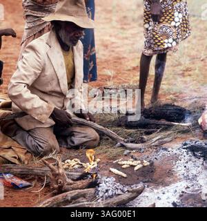 Gruppe von Aborigines im australischen Outback, die eine traditionelle Mahlzeit am offenen Feuer zubereiten. Ein älterer Mann mit Hut und abgenutztem Anzug kniet auf dem Boden und hält einen Stock, während Insektenlarven auf der heißen glut geröstet werden. Die Szene spielt sich in einer trockenen, rötlichen Landschaft ab, typisch für das australische Hinterland, um 1985. Banque D'Images