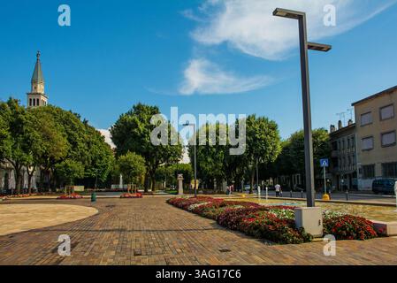 Piazza della Repubblica à Monfalcone, Frioul, Italie. Place avec fontaine, parterres de fleurs d'impatiens. A gauche : clocher de la cathédrale St Ambroise Banque D'Images