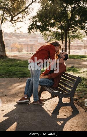 Moment romantique capturé dans un parc comme un couple partage une étreinte intime sur un banc. L'homme est assis tandis que la femme se penche sur, créant une tendresse et l Banque D'Images