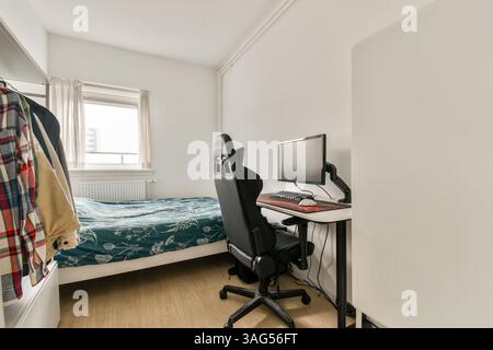 Un bureau à domicile bien rangé avec un bureau d'ordinateur, une chaise ergonomique et un lit confortable avec une couverture à motifs. La salle présente une lumière naturelle éclatante et des couleurs neutres. Banque D'Images
