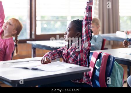 À l'école, fille afro-américaine levant la main tout en étant assise au bureau avec un cahier ouvert Banque D'Images