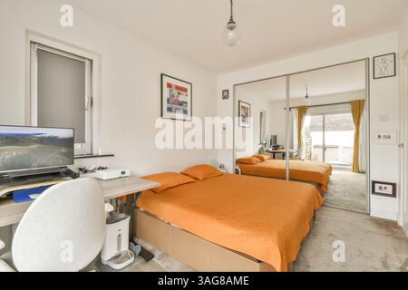 Cette chambre spacieuse et moderne dispose de lits jumeaux avec literie orange, d'un bureau fonctionnel et de grandes fenêtres qui offrent un éclairage naturel. Banque D'Images